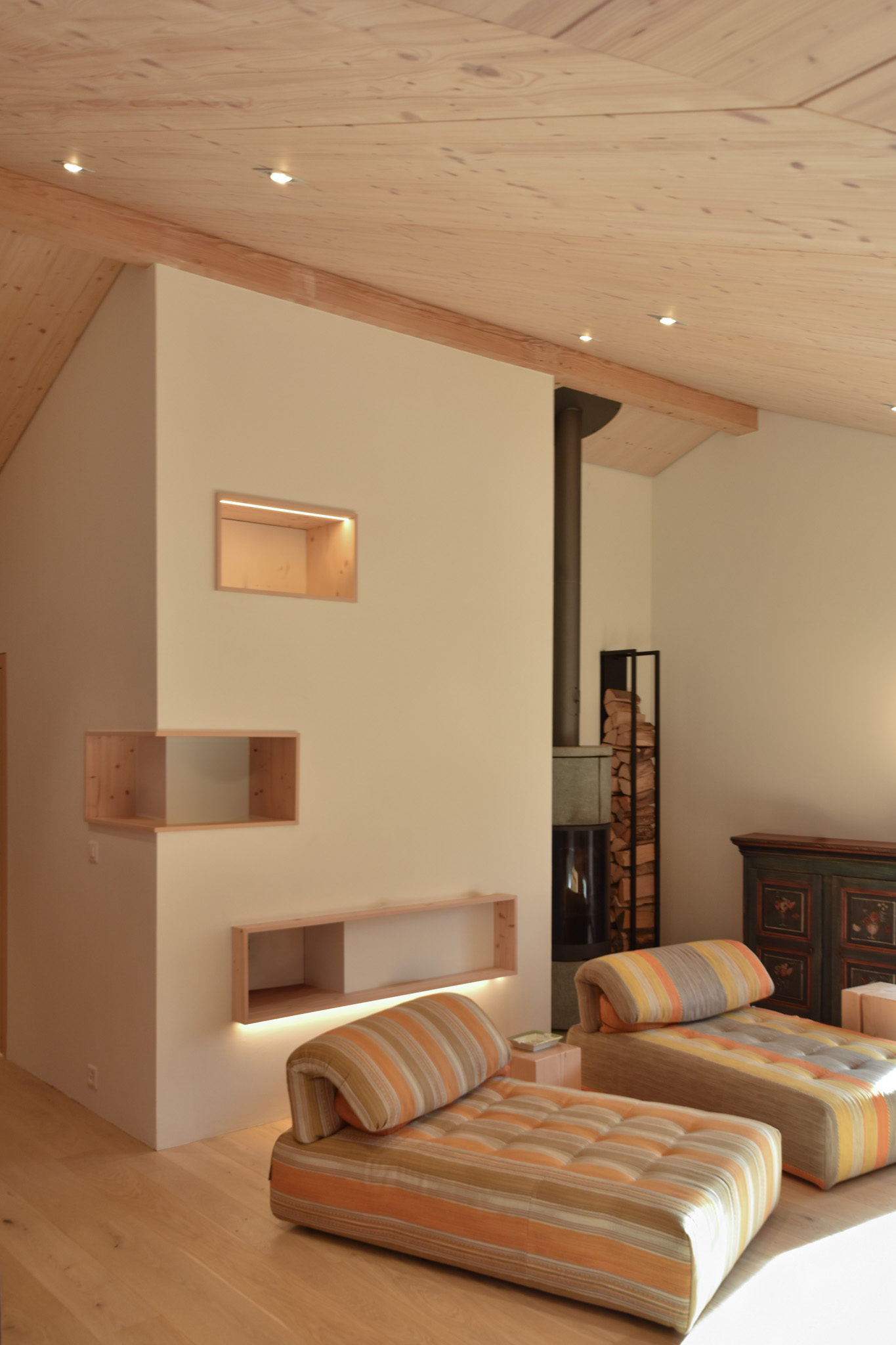 Cozy living room with two striped sofas, minimalist wooden shelves, a fireplace, and warm wooden ceilings. Natural light enhances the space.
