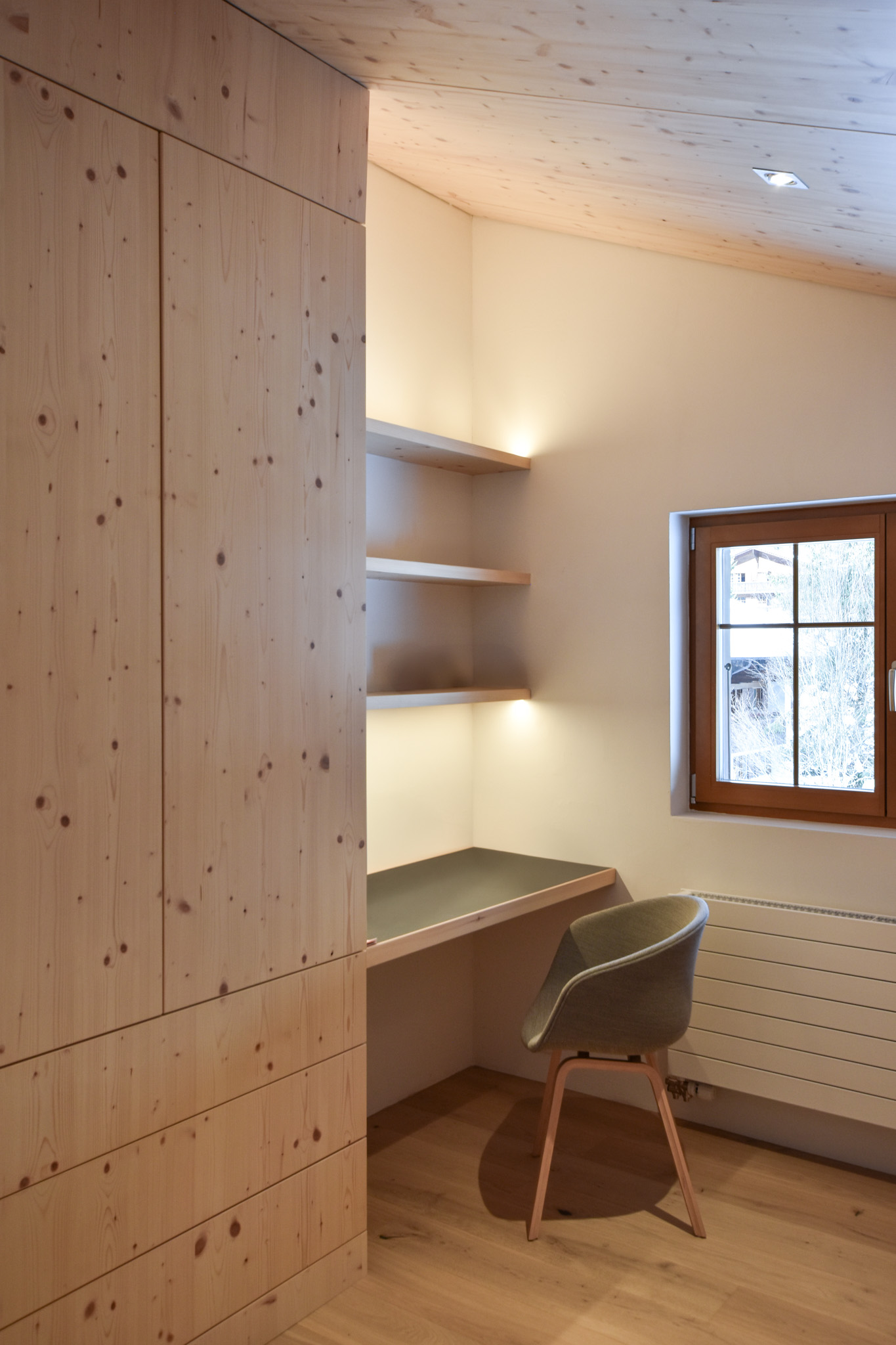 A cozy workspace featuring light wood storage, a simple desk, a soft chair, and a window letting in natural light.