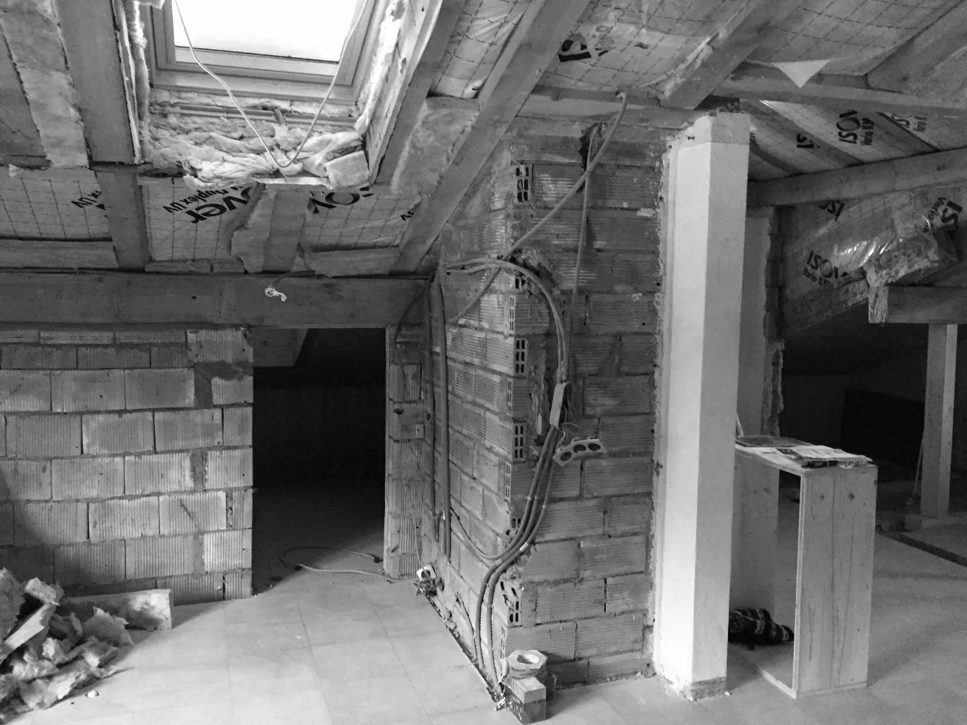 Black and white image of a partially finished attic with exposed brick walls, insulation, and a skylight above. Tools and debris scattered.