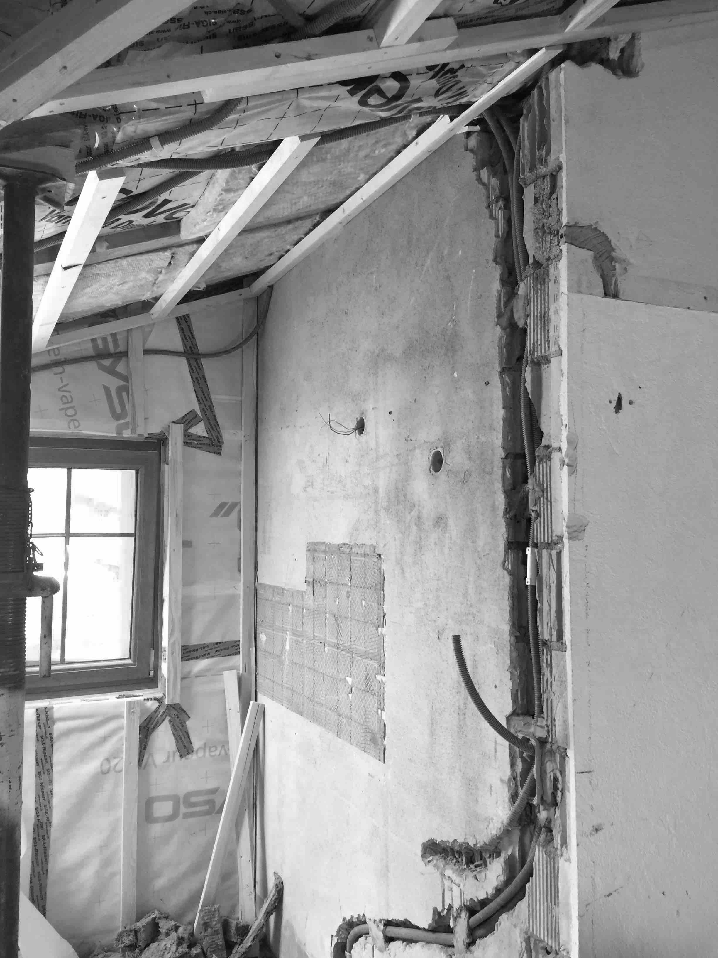 Black and white image of an under-renovation wall showing exposed wiring, insulation, and unfinished surfaces near a window.
