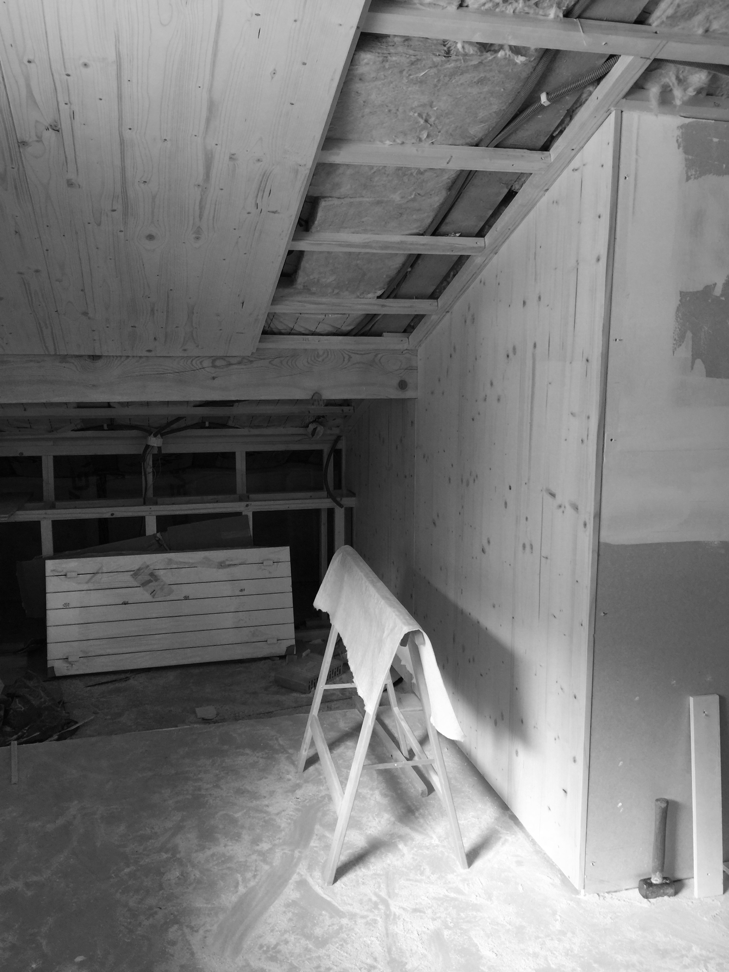 Building-site of an attic space featuring wooden beams, insulation, a sawhorse with a cloth, and construction debris on the floor.